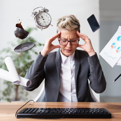 Image of a woman with hands to her temples with a phone, clock, spilling coffee cup and papers surrounding his head